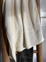 Et cremefarvet håndklæde er draperet over en håndklædestang af bambus med et mørkegråt håndklæde hængende under. Sollyset kaster bløde skygger, som fremhæver håndklædernes og bambussens organiske tekstur.